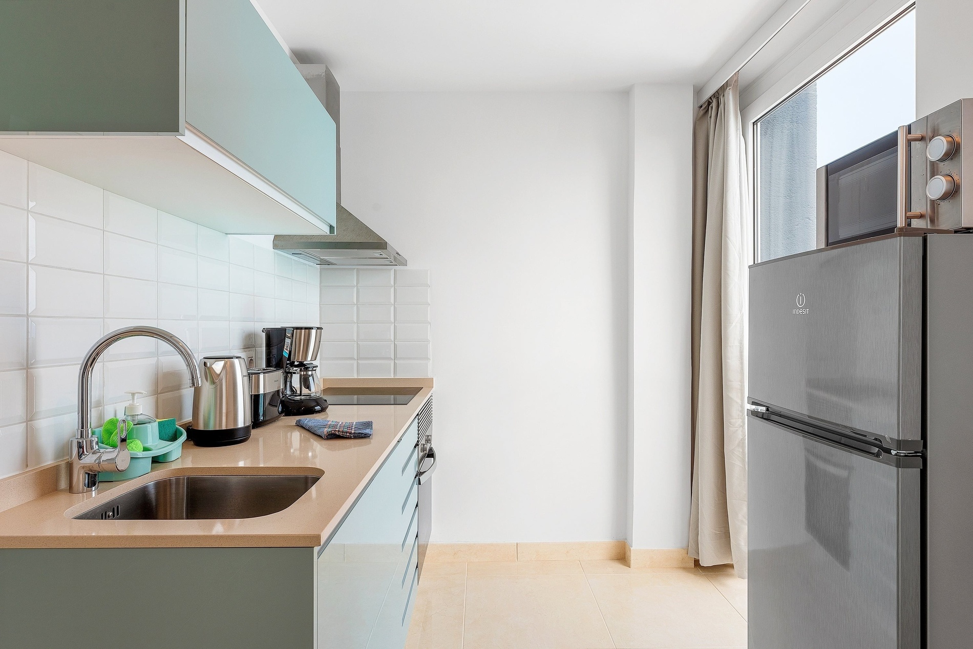 La photo présente une cuisine moderne et fonctionnelle, avec des meubles bleu-vert, un dosseret carrelé blanc, des appareils électroménagers en inox, un plan de travail beige et une fenêtre avec des rideaux clairs.