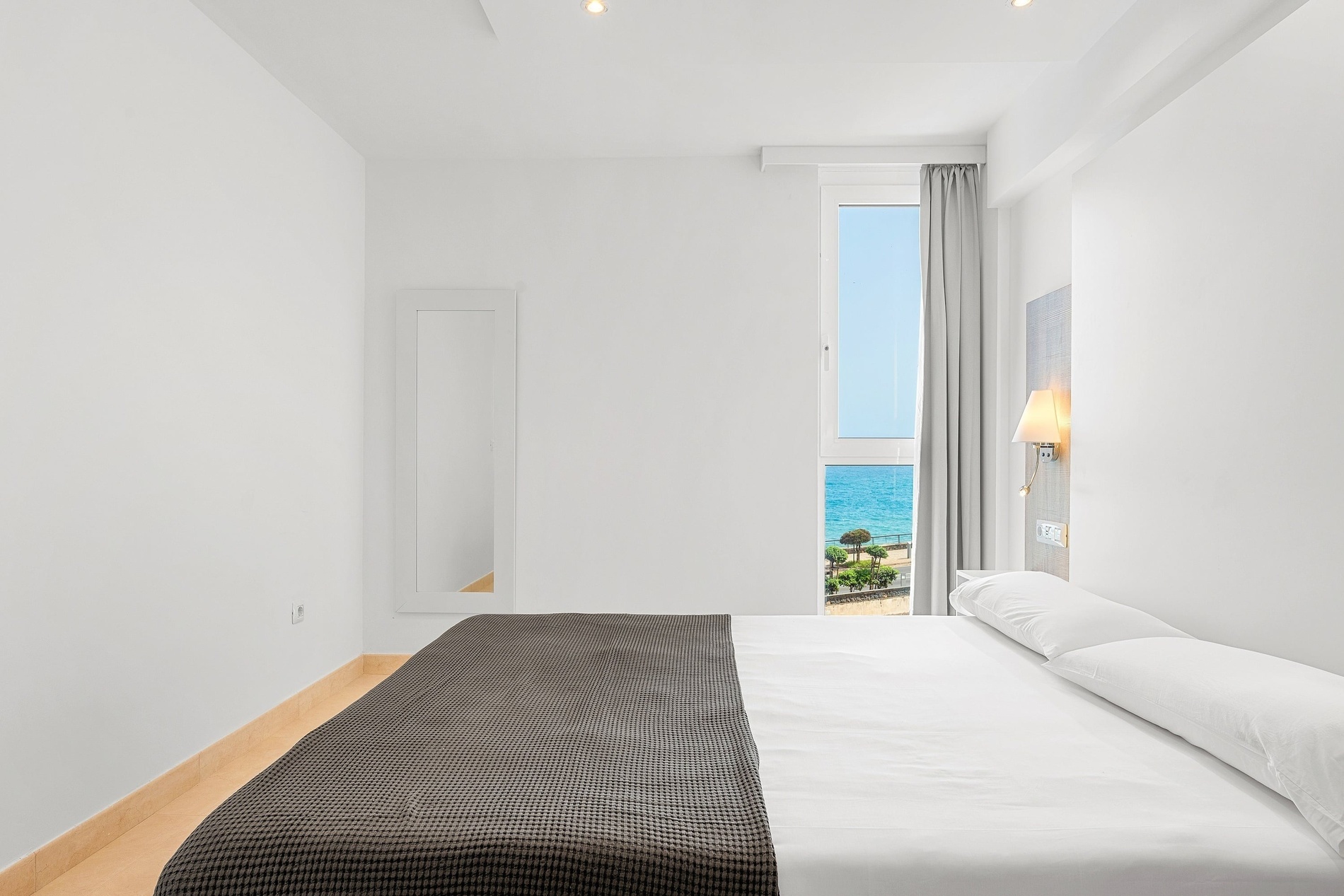 Une chambre lumineuse et minimaliste avec un grand lit, des murs blancs et une fenêtre verticale donnant sur la mer.