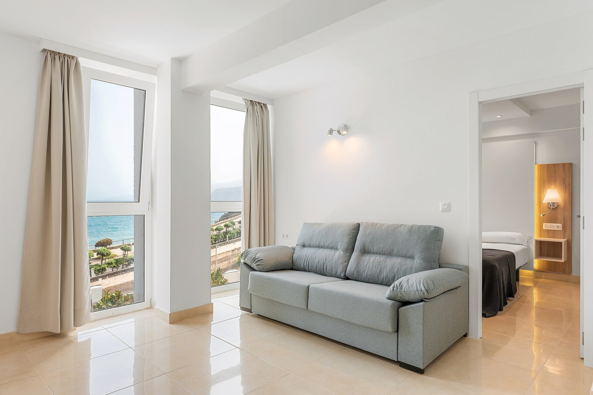 Une pièce lumineuse avec un canapé gris, deux grandes fenêtres offrant une vue sur la mer, et une porte ouverte menant à une chambre adjacente.