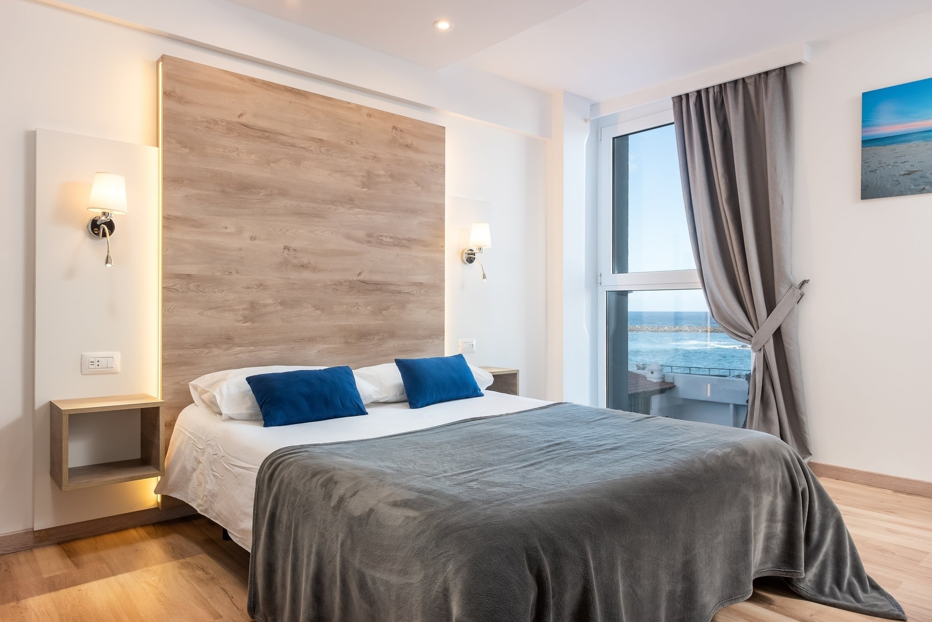 Une chambre moderne et lumineuse est présentée avec un grand lit orné de coussins bleus et d'une couverture grise, complété par une tête de lit en bois rétroéclairée et une grande fenêtre offrant une vue sur la mer.
