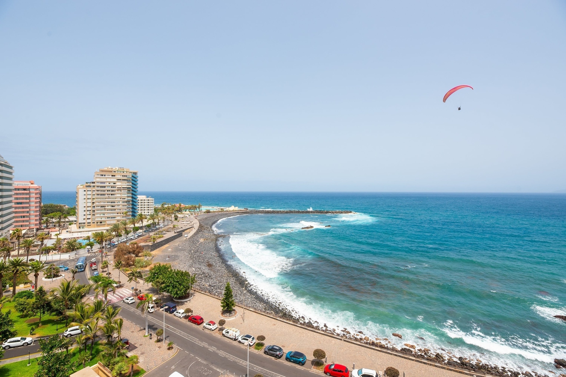 Une ville côtière animée avec des immeubles, des palmiers et des voitures le long de la plage de galets est visible sous un ciel bleu clair, tandis qu'un parapentiste plane au-dessus de l'océan turquoise.