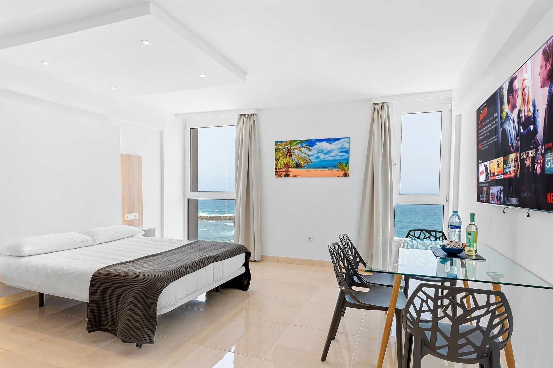 Un studio moderne et lumineux avec vue sur la mer, doté d'un lit, d'une table à manger en verre et d'un grand téléviseur mural.