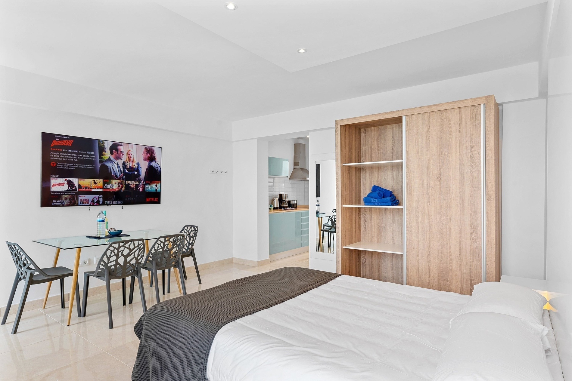 Cette image montre un studio moderne et lumineux, aménagé avec un lit, une table à manger avec des chaises, une télévision affichant Netflix, une kitchenette et une armoire.