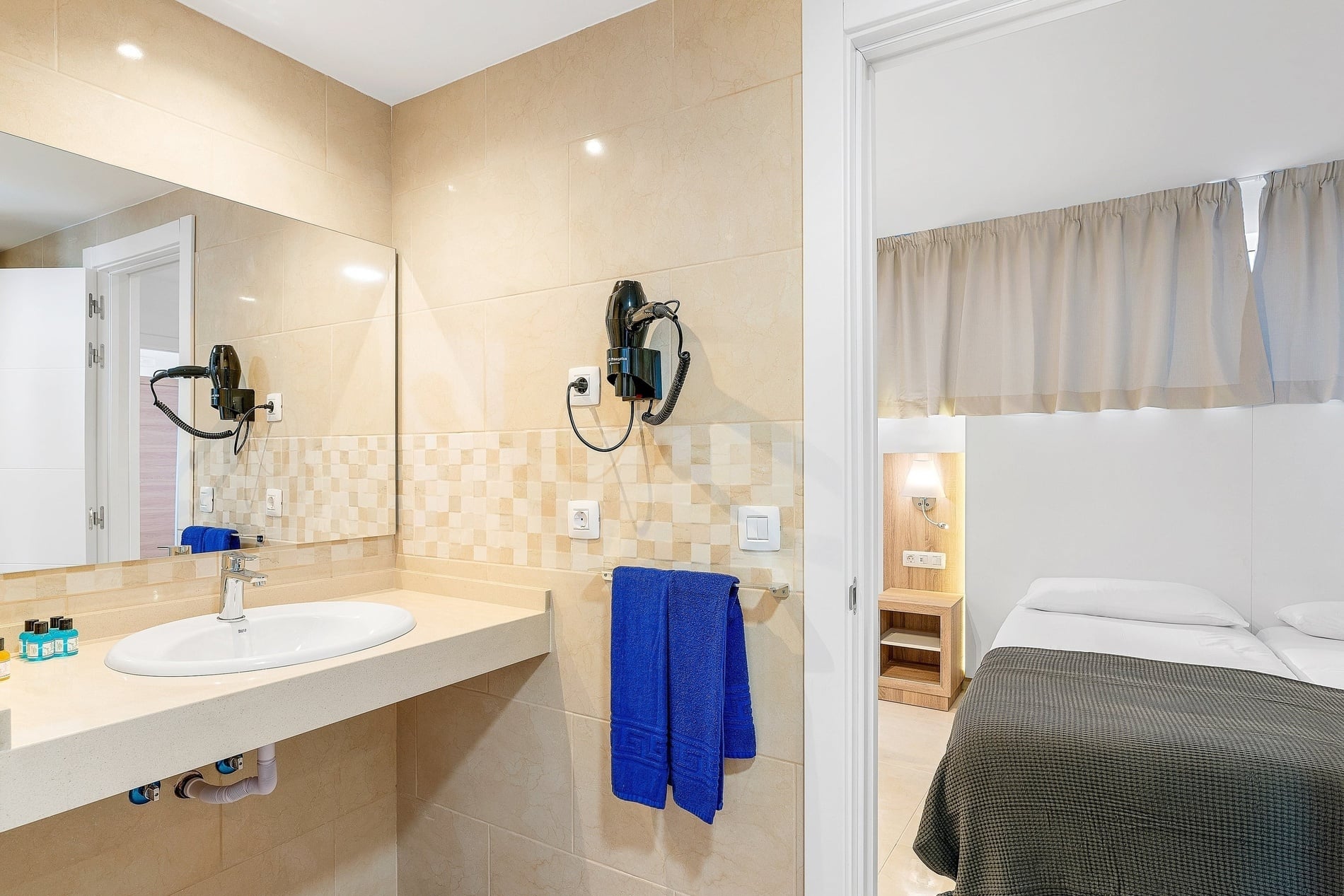 La photo montre une salle de bain carrelée avec un lavabo, un miroir et un sèche-cheveux, ouvrant sur une chambre à coucher avec un lit et une table de chevet.