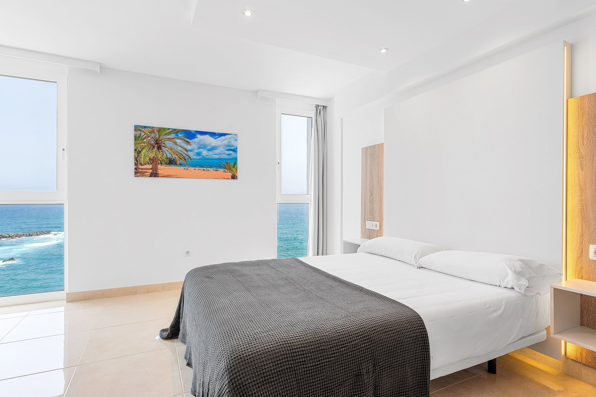 Cette chambre moderne et lumineuse offre une vue imprenable sur l'océan depuis sa grande fenêtre, avec un lit confortable, des murs blancs et un sol carrelé clair.