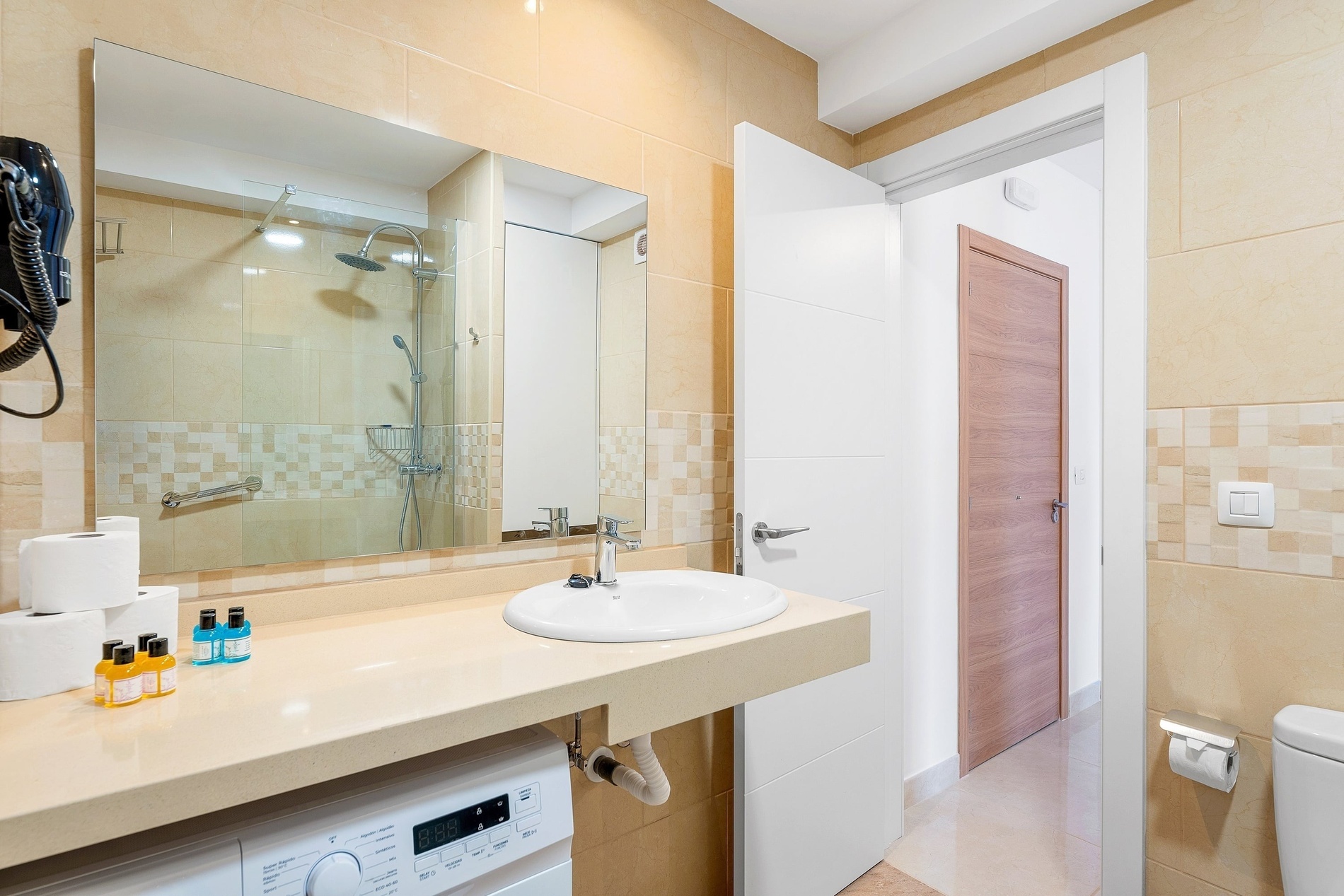 Une salle de bain moderne et fonctionnelle, aux murs carrelés beiges, équipée d'un lavabo avec un grand miroir reflétant la douche, et d'une machine à laver intégrée sous le plan de travail.