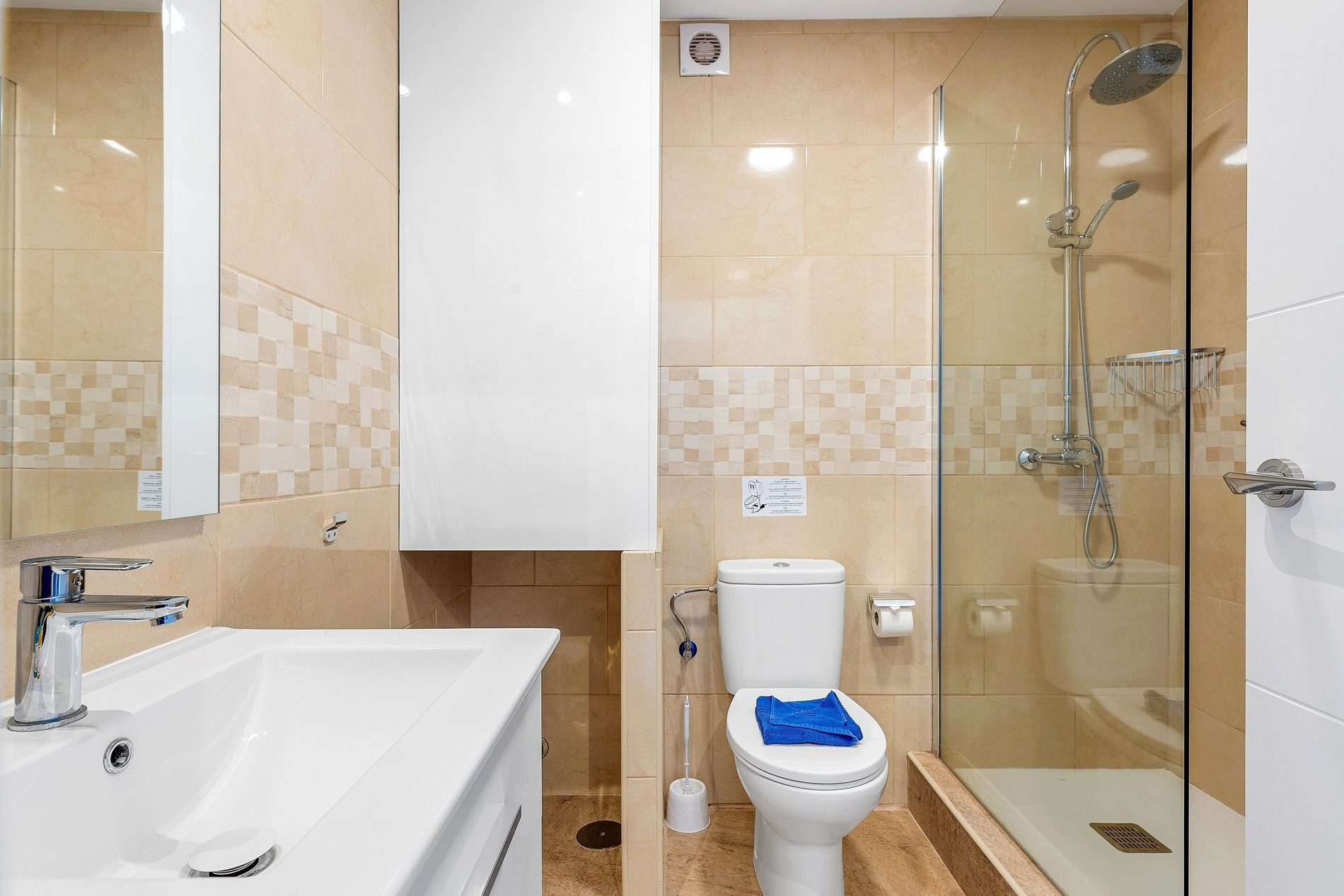 Cette image montre une salle de bain moderne aux murs carrelés de beige, équipée d'un lavabo blanc, de toilettes sur lesquelles repose une serviette bleue, et d'une douche avec paroi vitrée.