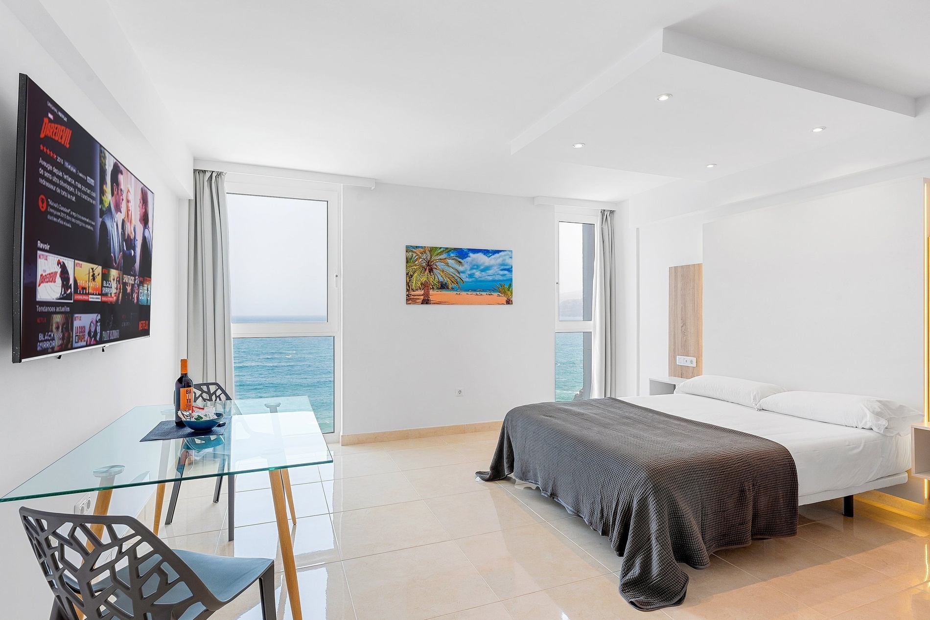 Une chambre moderne et lumineuse offre une vue imprenable sur la mer, avec un lit, une télévision diffusant Netflix, et une table en verre avec des chaises.