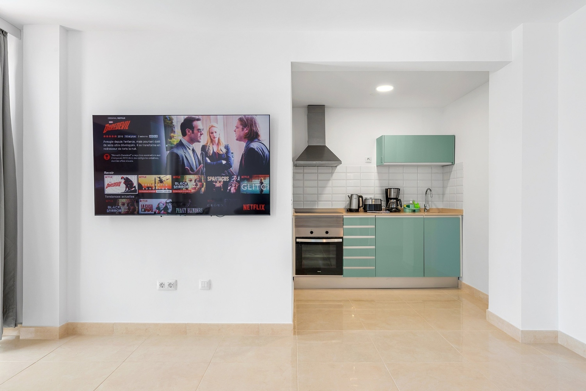 L'image montre un espace de vie moderne avec une télévision murale affichant Netflix et une cuisine ouverte aux armoires vert menthe et aux appareils électroménagers en acier inoxydable.