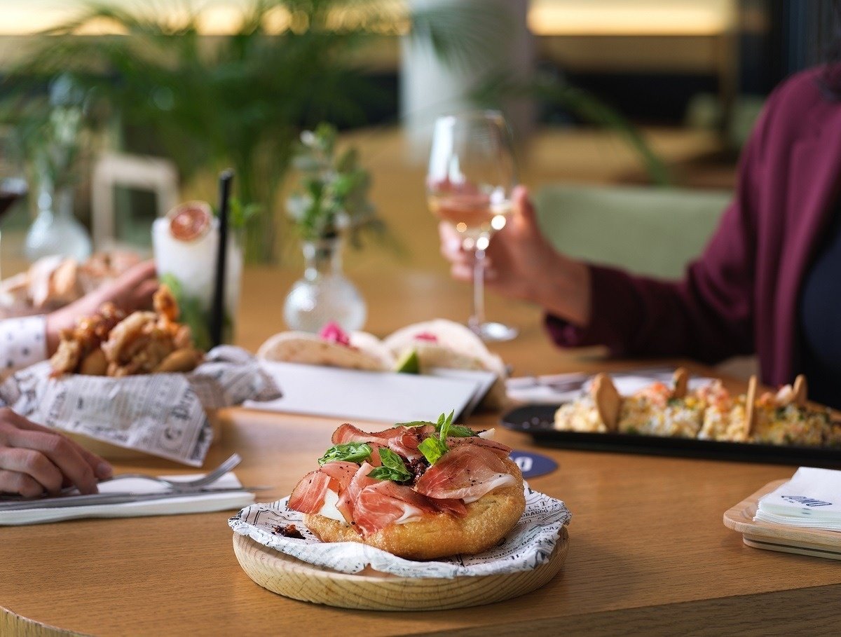 a person holding a glass of wine sits at a table with food