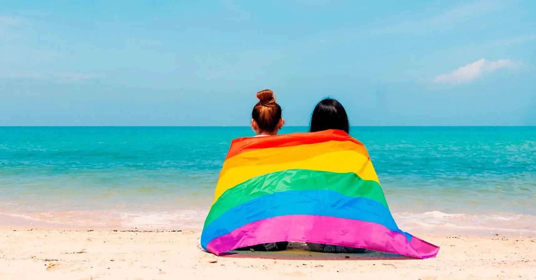 dos mujeres están sentadas en la playa con una bandera del arco iris
