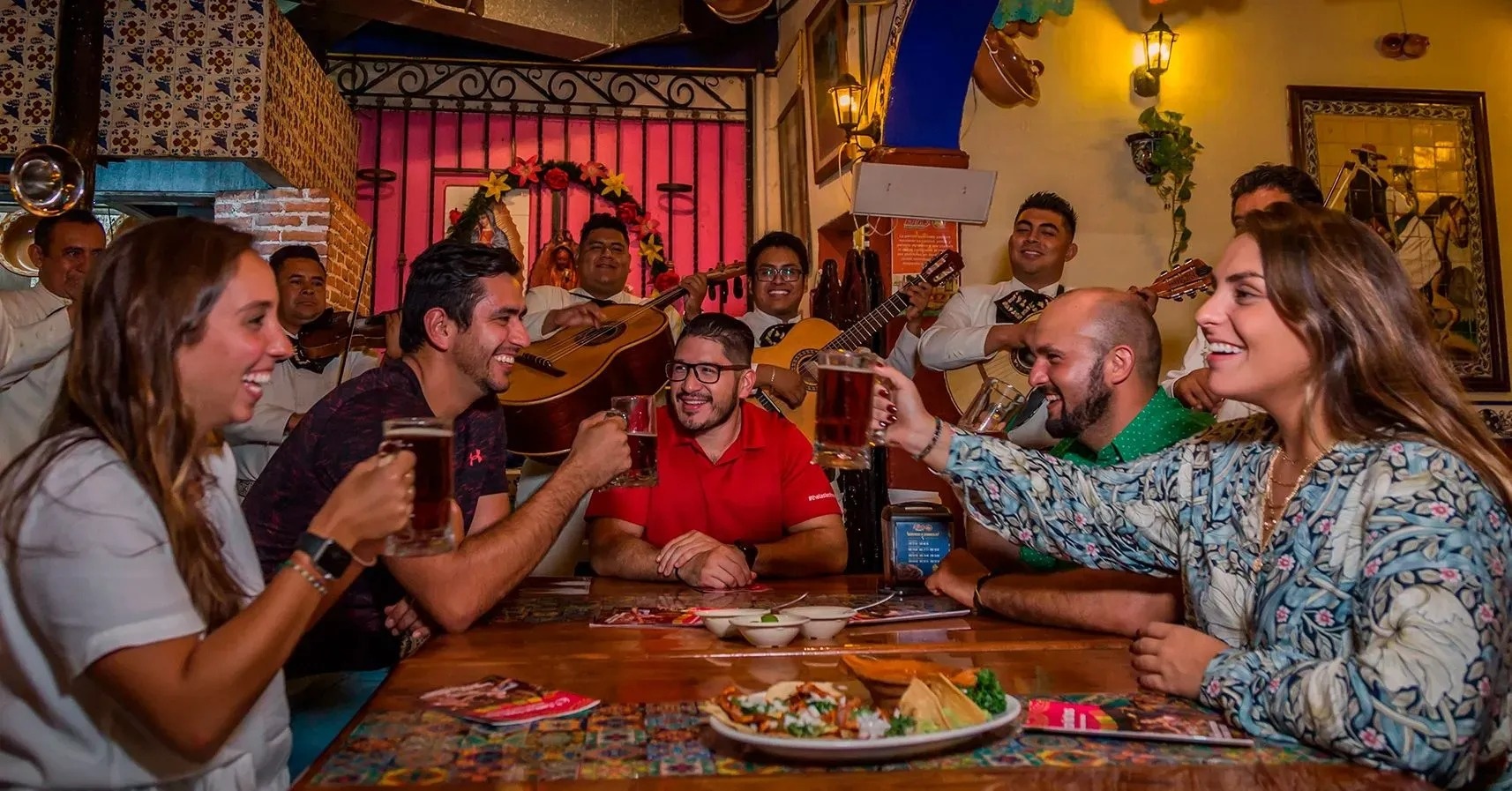 un grupo de hombres y mujeres se sientan alrededor de una mesa comiendo tacos y bebiendo cerveza