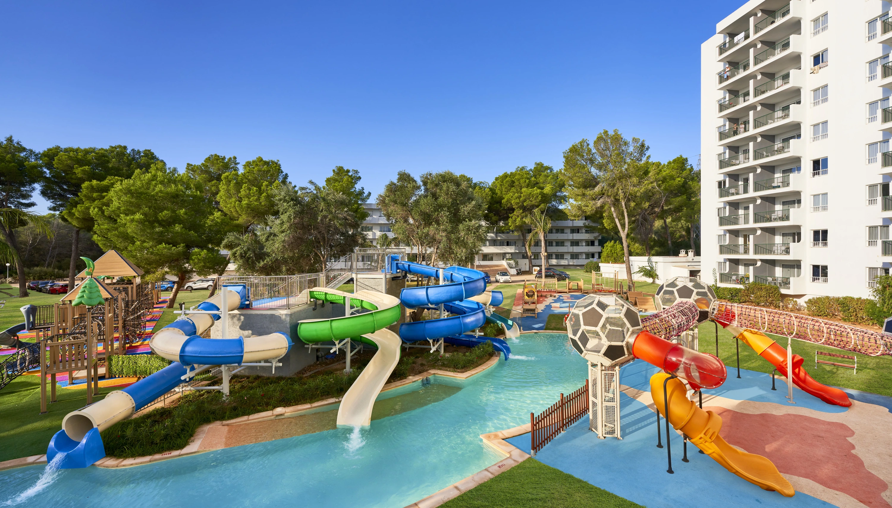 Una vista panoràmica d'un complex turístic amb una gran piscina, un parc aquàtic, edificis d'apartaments blancs i una muntanya àrida al fons, sota un cel blau clar.