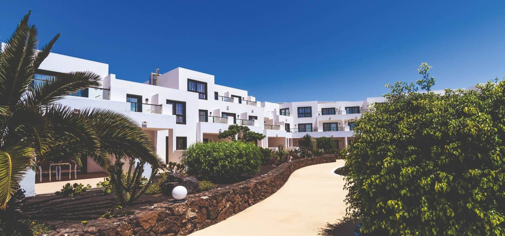BlueBay Lanzarote | O7 Hotels | Illes Canàries: hotel blanc amb palmeres i piscina infinita sota cel blau.