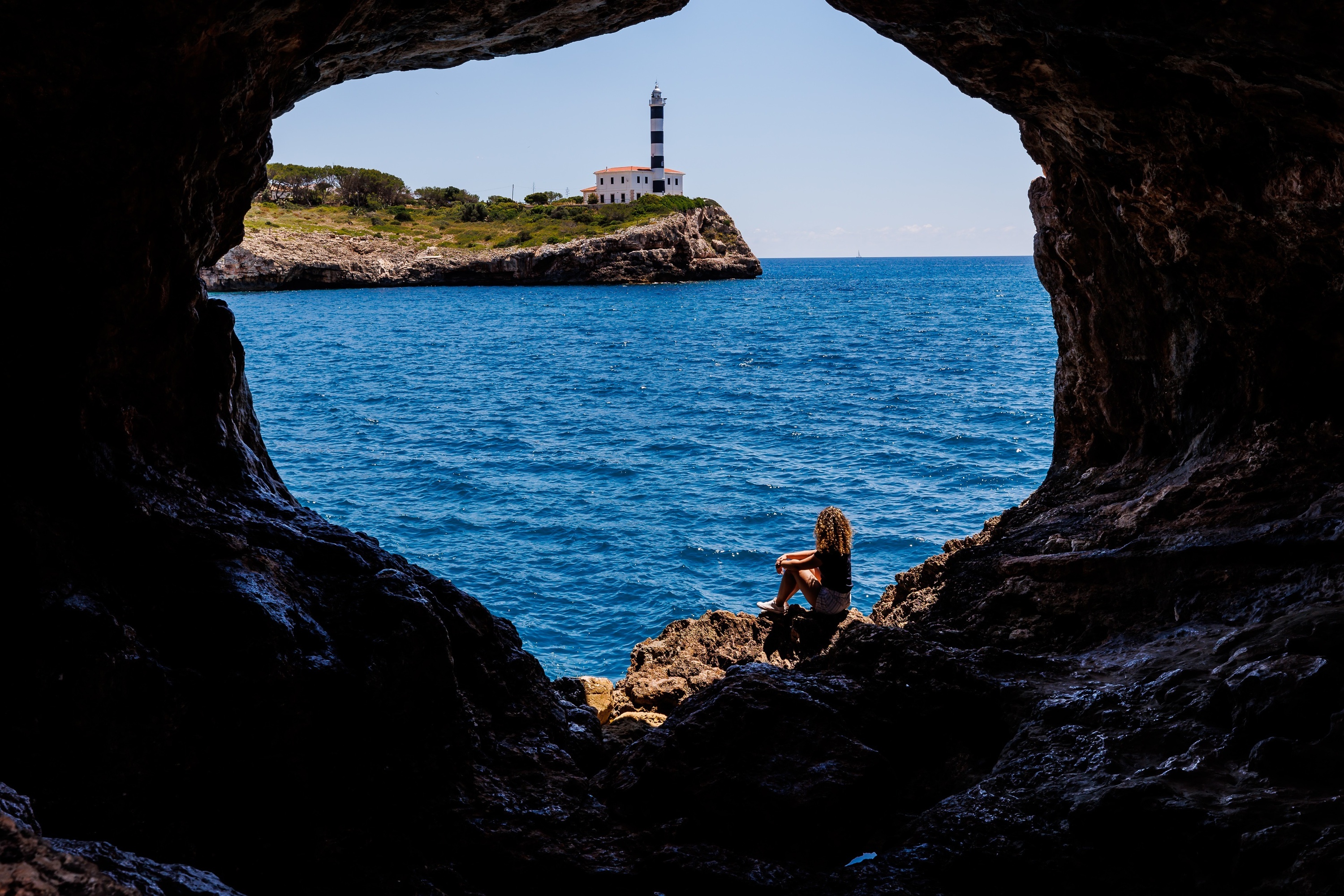 una donna si siede in una grotta con vista sull'oceano e un faro sullo sfondo