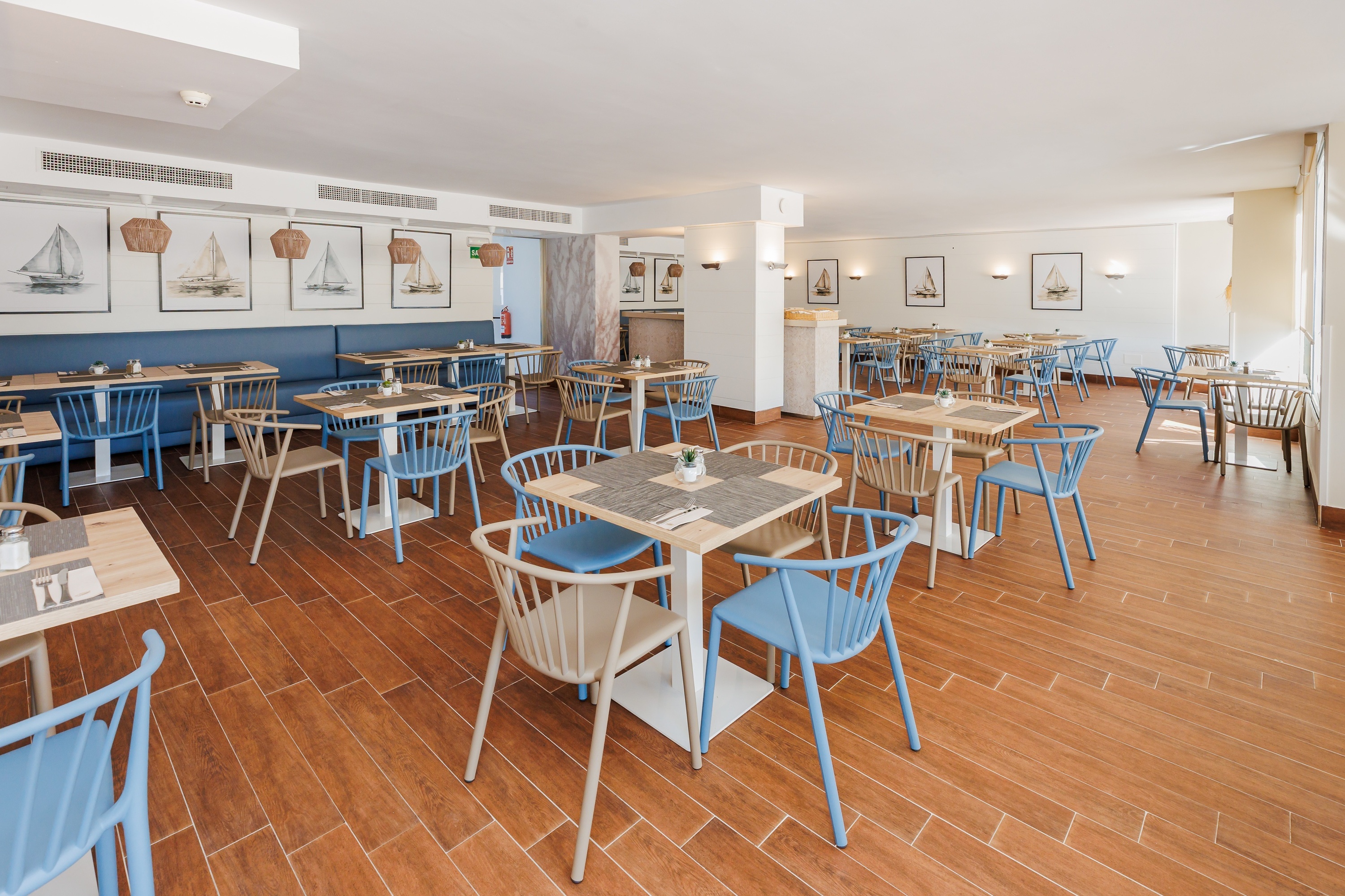 Un espaiós i lluminós restaurant modern amb decoració nàutica, cadires blaves i beix, i terra d'imitació fusta.