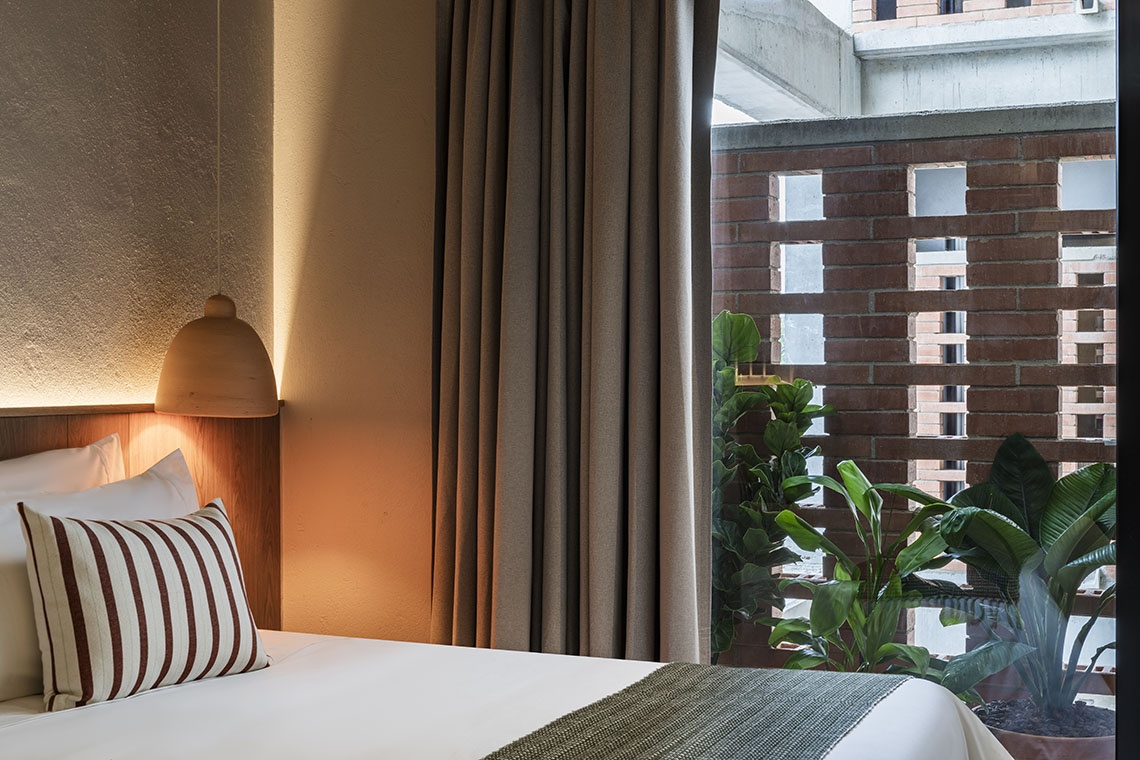 Bedroom at Obra Hotels Medellin with hanging lamp, striped pillow, and greenery visible through brick window.