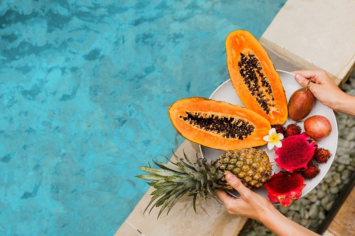 una persona sostiene un plato de frutas tropicales junto a una piscina