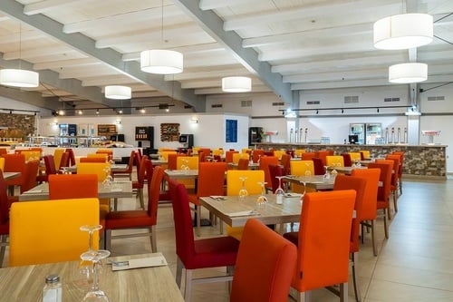 Un amplio y luminoso comedor de restaurante se presenta con numerosas mesas dispuestas y sillas de colores vibrantes como rojo, naranja y amarillo, mientras que al fondo se aprecian estaciones de buffet y un área de bar.