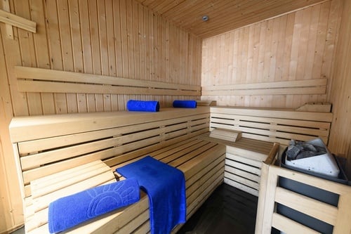 Voici l'intérieur chaleureux d'un sauna en bois clair, équipé de bancs à plusieurs niveaux, de quelques serviettes bleues pliées et d'un poêle de sauna rempli de pierres dans le coin droit.