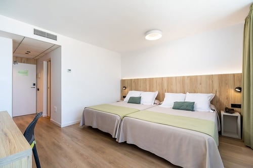 Una habitación de hotel moderna y minimalista con dos camas individuales, paredes blancas, cabecero de madera clara y detalles en verde menta.