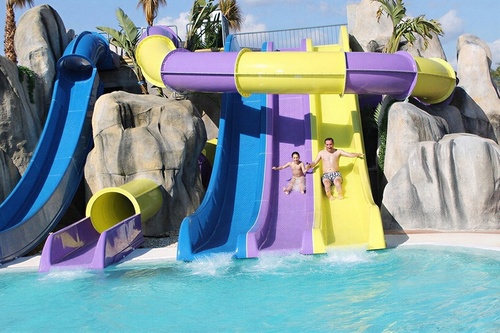 An adult and a child slide down colorful water slides into a pool at a water park, surrounded by artificial rocks and palm trees.