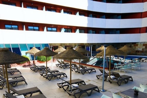Vista de un complejo hotelero moderno con fachada de balcones, una piscina y una terraza con tumbonas y sombrillas de paja.