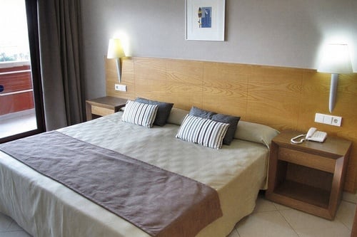 Un dormitorio de hotel con una cama hecha, dos mesitas de noche con lámparas de pared y una puerta de cristal que conduce a un balcón.