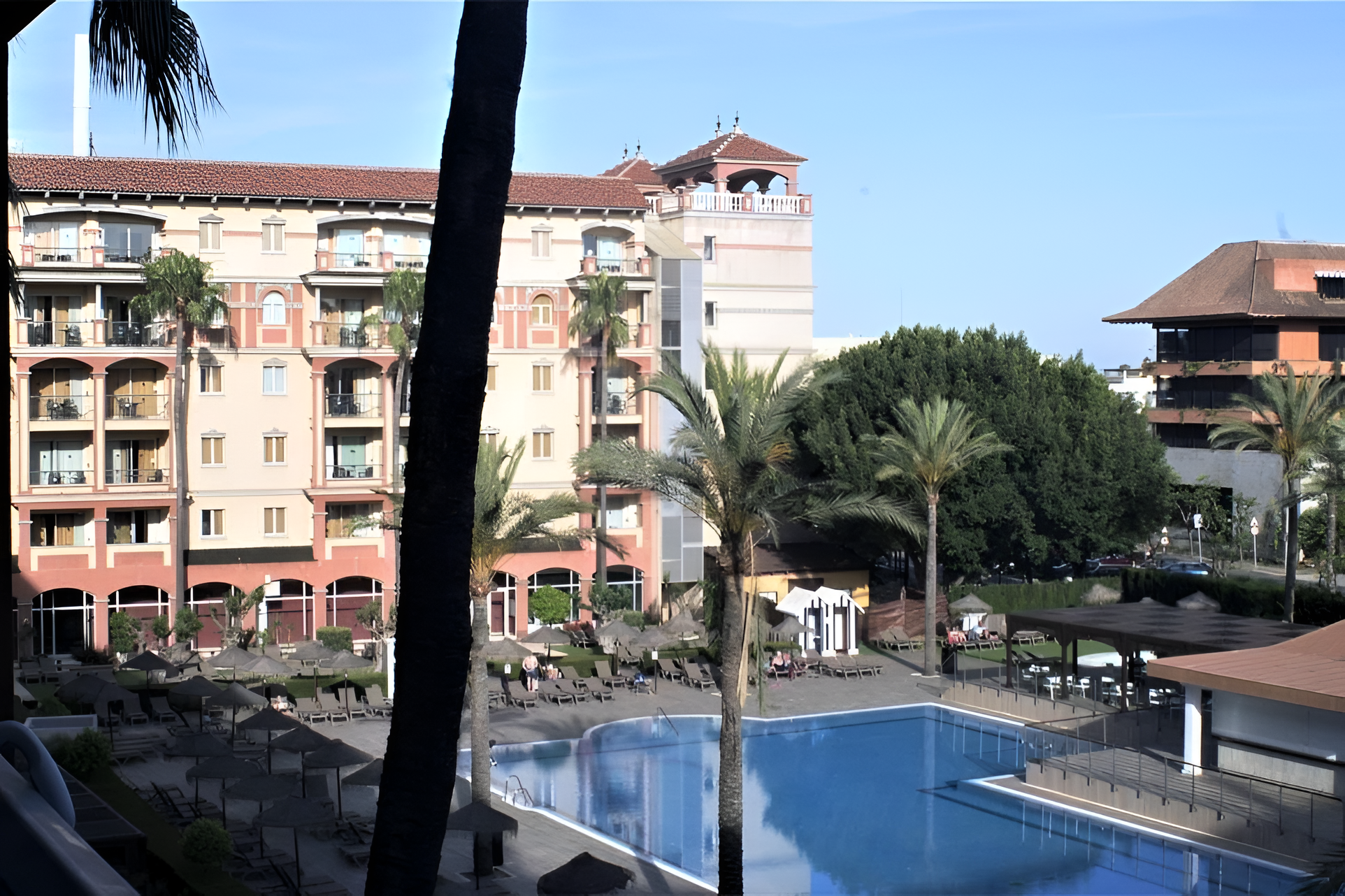 La imagen muestra un gran hotel con balcones, palmeras, una piscina con tumbonas y sombrillas, y otro edificio con mucha vegetación de fondo.