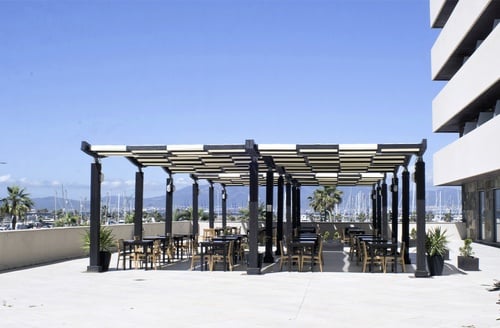 Hotel patio with tables under a pergola, overlooking a marina in La Linea de la Concepcion.