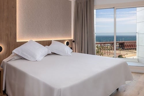 Chambre d'hôtel moderne et lumineuse avec un lit blanc immaculé et une grande fenêtre donnant sur la mer, un viaduc métallique et des palmiers.
