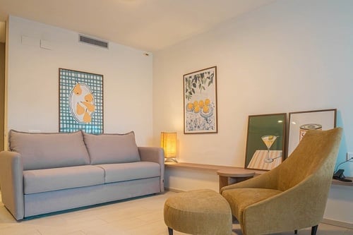 Una sala de estar moderna y luminosa con un sofá gris claro, un sillón amarillo con otomana y varias obras de arte en las paredes blancas.