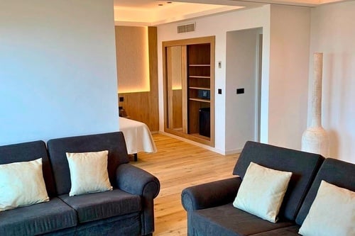 L'image présente une suite d'hôtel moderne et lumineuse, avec un coin salon doté de canapés foncés et une chambre à coucher visible à travers une ouverture, le tout agrémenté de boiseries claires.