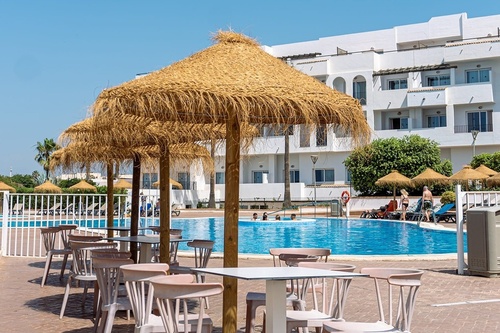 A resort pool area is bustling with activity under a clear sky, featuring straw umbrellas shading tables and chairs in the foreground, and a white hotel building in the background.