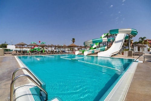 Una vista amplia de una piscina de hotel con toboganes de agua coloridos, tumbonas y sombrillas de paja bajo un cielo azul despejado.
