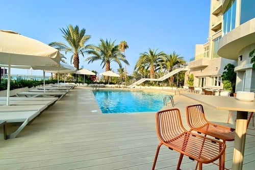 Une piscine extérieure bordée de palmiers, de chaises longues et de parasols est visible devant un bâtiment moderne sous un ciel clair et ensoleillé.