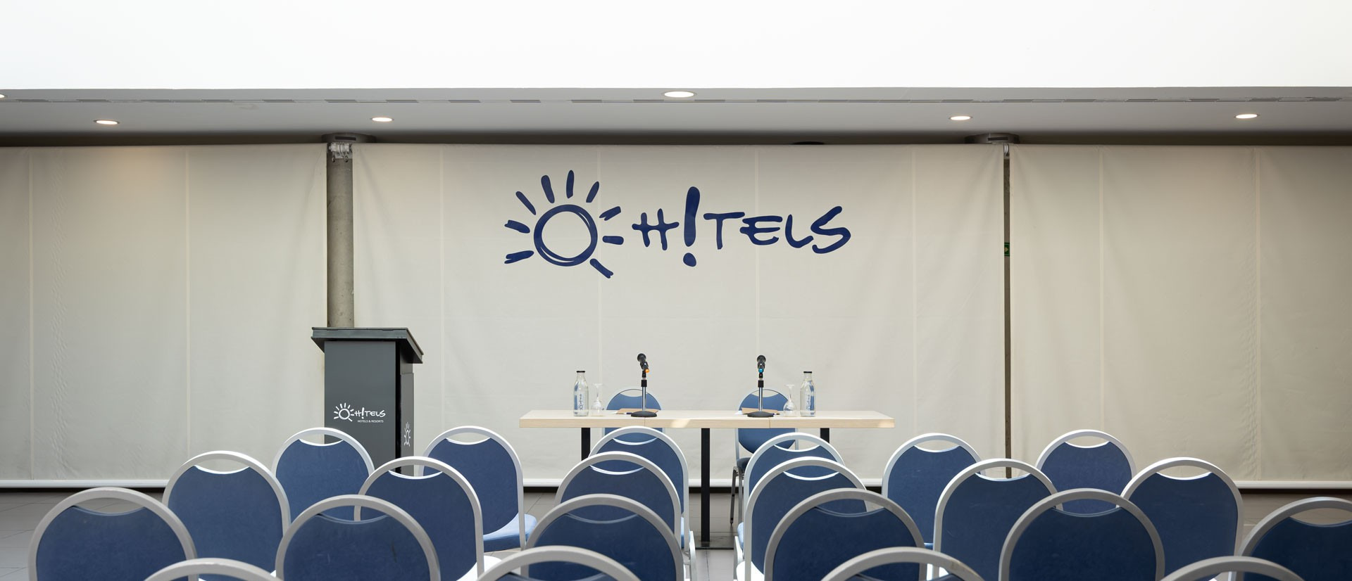 Sala de conferencias con logo de Organiza tu evento | Ohtels Hotels & Resorts | Web oficial, atril y sillas azules.