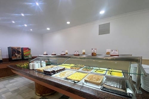 Una mesa de buffet con una variedad de postres, frutas y dispensadores de bebidas en un comedor bien iluminado.