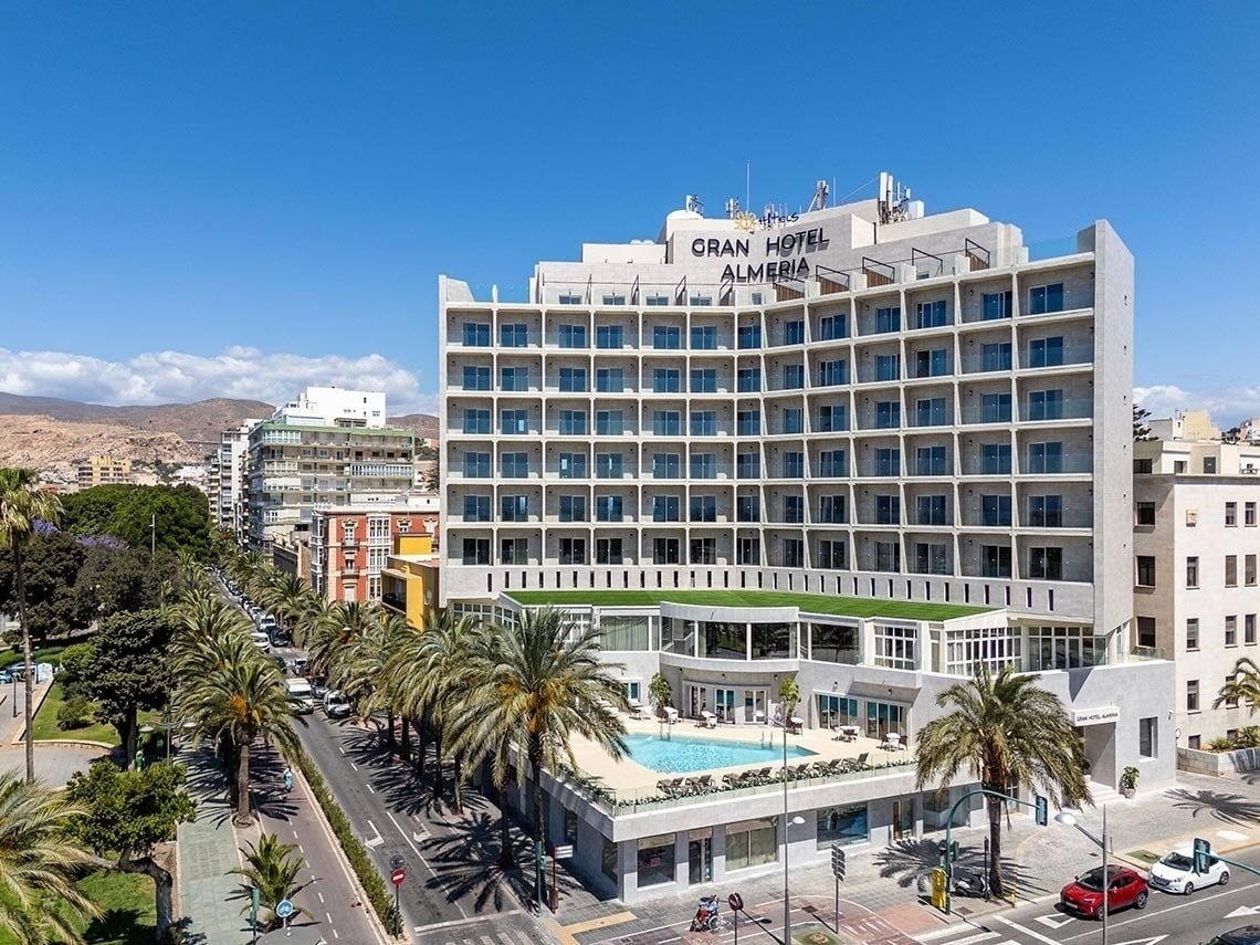 The entrance of the Gran Hotel Almeria is shown with its name prominently displayed above glass doors, flanked by potted plants and set against a light grey tiled facade.
