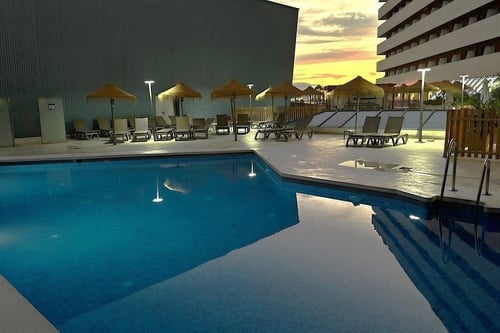 Una piscina exterior de hotel con sombrillas y tumbonas se ilumina al atardecer, reflejando el cielo y los edificios circundantes en sus aguas.