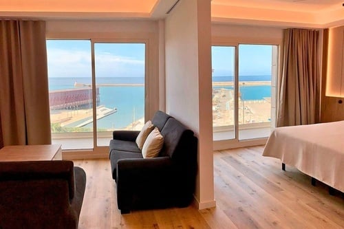 Une chambre d'hôtel moderne et lumineuse avec de grandes baies vitrées offrant une vue imprenable sur un port et la mer, comprenant un coin salon et un espace nuit séparés.