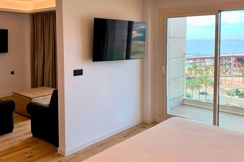 L'image montre un intérieur moderne avec un lit, un coin salon et une télévision, et une grande fenêtre donnant sur la mer, un pont et des palmiers.