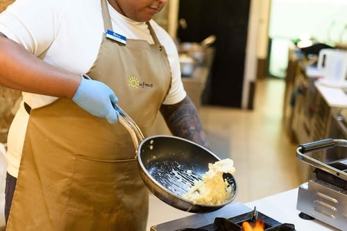 Un cuisinier, vêtu d'un tablier et de gants bleus, prépare des œufs brouillés dans une poêle au-dessus d'une flamme.