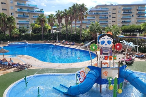 Un complejo de piscinas vibrante presenta una gran piscina, un parque acuático infantil con toboganes y una figura de calavera decorada, flanqueado por edificios y palmeras.
