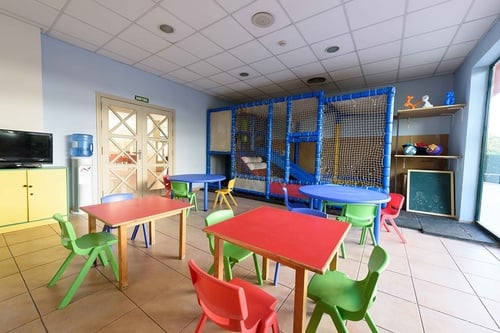 Une aire de jeux intérieure colorée pour enfants est équipée de petites tables, de chaises, d'une structure de jeu et d'une télévision.