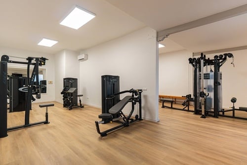 Este gimnasio moderno presenta un suelo de madera clara, paredes blancas y varias máquinas de ejercicio de color negro distribuidas en un espacio bien iluminado.