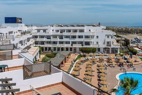 An elevated view captures a sprawling white resort complex with multiple buildings, a swimming pool surrounded by sun loungers and straw umbrellas, and distant mountains and coastline under a clear sky.