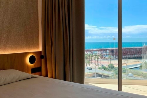 Chambre d'hôtel moderne avec un lit, un éclairage d'ambiance et une grande fenêtre offrant une vue sur la mer, une jetée et des palmiers.