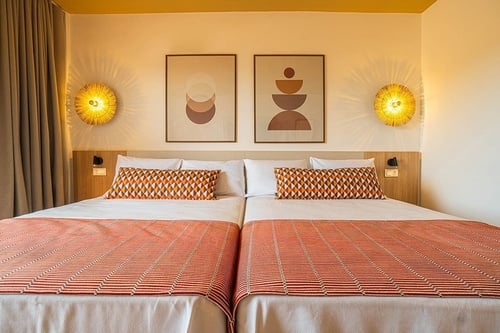 Un dormitorio de hotel con dos camas individuales, decorado con arte abstracto, lámparas de pared de fibras naturales y textiles geométricos en una paleta de colores cálidos.