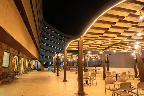 Un patio exterior iluminado con una pérgola y mobiliario de terraza se extiende frente a un moderno edificio curvo con balcones, todo bajo el cielo nocturno.